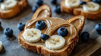 Creative breakfast toast shaped as cat face with peanut butter, banana slices as eyes, and blueberry nose on rustic wooden background. Fun food art for kids.