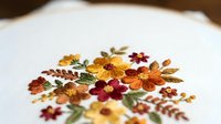 Delicate autumn floral embroidery on white fabric featuring orange, burgundy and yellow flowers with green leaves in traditional needlework style.