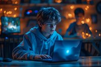 Teenage boy in white hoodie using laptop at night, illuminated by blue screen light and warm bokeh background. Moody atmospheric home setting.