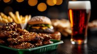 Juicy chicken wings with sesame seeds served alongside classic cheeseburgers and french fries. Dark restaurant setting with amber bokeh lights and craft beer glass.