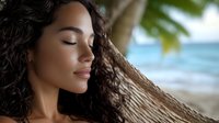 Woman with long curly dark hair and medium skin tone relaxing against palm tree trunk on tropical beach with ocean and blue sky in background, eyes closed in peaceful moment.