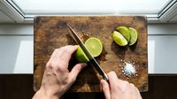 Chef's hands holding knife and lime on wooden cutting board with sea salt and chopsticks for cooking, food preparation, and culinary design projects.