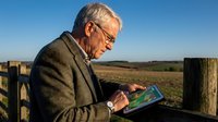 Senior Caucasian businessman in suit using tablet outdoors by wooden fence with rural landscape background for technology and agriculture concepts.