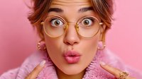 Young woman in round glasses making surprised expression with puckered lips against pink background, wearing fuzzy pink sweater and gold accessories.