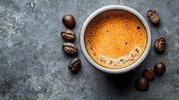 Fresh espresso with golden crema in white ceramic cup surrounded by roasted coffee beans on dark textured background, top view food photography.