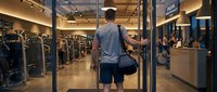 Athletic man entering modern fitness gym with sports bag, ready for workout session in well-equipped training facility with exercise machines and weights.