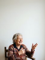 Senior woman with gray hair and fair complexion, wearing a rust-colored patterned cardigan, sitting on a wooden chair against a white background, smiling and gesturing expressively while speaking into a microphone for presentation or interview purposes.