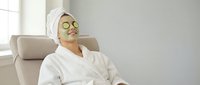 Young Asian woman relaxing in white bathrobe with cucumber eye mask on comfortable chair, enjoying spa treatment and self-care routine at home.