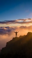 Silhouette of person with raised arms celebrating success on mountain peak above clouds during golden hour sunrise with dramatic sky for motivation concepts.