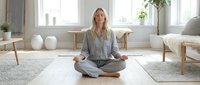 Woman in casual gray clothing sitting in lotus position on yoga mat in bright, minimalist living room with natural light, practicing meditation and mindfulness for wellness and relaxation.