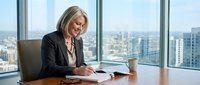 Mature blonde woman in business attire writing at desk in high-rise office with city skyline view through floor-to-ceiling windows, professional corporate environment.