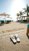 White sandals on a woven mat at a luxury beach resort, word vacation written in sand, palm trees and lounge chairs in background.