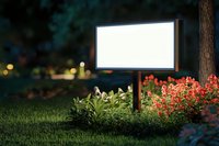 Illuminated blank billboard in garden setting with red flowers and green grass at dusk, mockup for outdoor advertising display against blurred bokeh background.