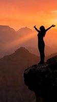 Woman silhouette with raised arms celebrating freedom on mountain cliff at golden sunset with layered peaks and orange sky for wellness and adventure content.