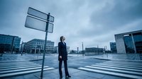 Professional businessman in dark suit standing at crossroads with blank directional signs in modern urban plaza during overcast day symbolizing business decisions.