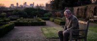 Senior Caucasian man sitting alone on wooden bench in urban park during golden hour sunset with city skyline background for loneliness and retirement concepts.