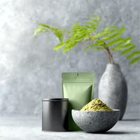 Green matcha tea powder in stone bowl with packaging and decorative fern leaf in ceramic vase against concrete wall background, minimalist composition.