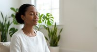 Young African American woman practicing mindfulness meditation at home with eyes closed, sitting peacefully near green plants in bright natural light.