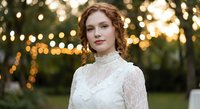 Young Caucasian woman in elegant white lace dress with curly red hair posing outdoors with warm bokeh lights background for wedding or formal event photography.