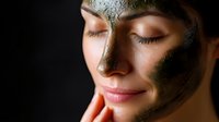 Young woman with clay face mask during spa treatment, eyes closed in relaxation against dark background, showing skincare and wellness routine.