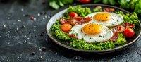 Sunny side up eggs served on fresh green salad with crispy bacon strips, cherry tomatoes and herbs on rustic plate against dark stone background.