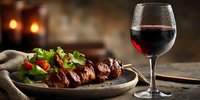 Grilled meat skewers with fresh salad and red wine glass on rustic wooden table in cozy restaurant setting with warm ambient lighting.