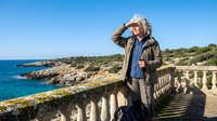 Mature blonde woman enjoying coastal scenery from stone balustrade overlooking Mediterranean sea and rocky shoreline on sunny day.