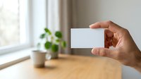 Hand holding blank white business card in modern office workspace with potted plant and natural window light for branding mockup.