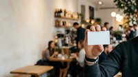 Unrecognizable male hand holding blank white card at festive gathering with decorated interior and blurred guests in background for promotional messaging.