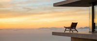 Solitary lounge chair on modern cantilever deck overlooking misty mountain landscape at golden sunset, conveying luxury relaxation and serene escape.