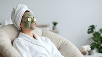 Young Caucasian woman in white robe relaxing with green clay face mask and cucumber slices, enjoying home spa skincare routine.