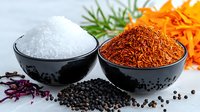 Aromatic spices and seasonings in black ceramic bowls - sea salt, saffron threads, and black peppercorns on white surface with fresh herbs and flowers.