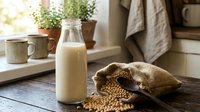 Fresh soy milk in glass bottle with scattered soybeans on rustic wooden table in cozy kitchen setting with natural morning light and plants.