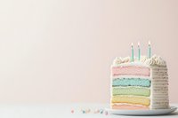 Pastel rainbow layer cake with white frosting and lit birthday candles on white plate against soft pink background, decorated with colorful sprinkles.