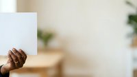 Person of African descent holding blank white card or paper in front of face in bright indoor space with blurred background, suitable for advertising, marketing campaigns, and promotional materials.