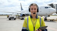 Female airport ground crew worker with headset smiling at camera wearing high visibility vest on tarmac with commercial aircraft in background at busy airport.