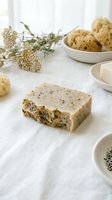Handmade herbal soap bar with dried botanicals and natural ingredients displayed on white linen with fresh flowers, cookies, and spa accessories for wellness and beauty product marketing.