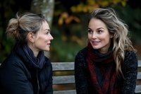 Two young Caucasian women having friendly conversation on park bench wearing cozy autumn scarves and sweaters during fall season outdoors.
