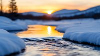 Tranquil winter stream with golden sunset reflection between snowy banks, silhouetted mountains and evergreen trees create serene wilderness landscape.