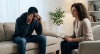 African American man experiencing stress during therapy session with female counselor in modern office setting.