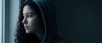 Young woman with curly hair in hoodie looking pensively out window in moody lighting for mental health awareness campaigns and emotional storytelling.