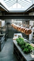 Professional kitchen interior with hanging copper pots, stainless steel appliances, skylight ceiling, and fresh herbs on a concrete island.