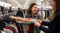Young adult female shoppers browsing colorful clothing on hangers in retail store, one wearing patterned jacket smiling while selecting garments, busy shopping environment with customers and merchandise.