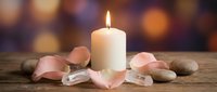 Burning white candle surrounded by pink rose petals and smooth stones on rustic wooden surface with warm bokeh lights background for spa relaxation meditation.