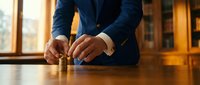 Professional businessman in navy suit stacking wooden blocks on polished table in warm office setting, representing strategic planning and business growth concepts.