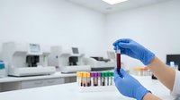 Medical laboratory technician in blue gloves holding blood sample test tube in modern clinical lab with automated equipment for diagnostic testing.