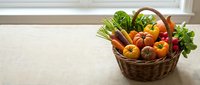 Wicker basket filled with fresh colorful vegetables including tomatoes, carrots, peppers, and greens on a linen surface near a window.