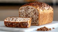 Fresh baked whole grain bread loaf with sliced piece and scattered flax seeds on white marble surface, warm natural lighting shows detailed texture.