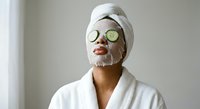 Young woman in white bathrobe with facial mask and cucumber slices on eyes enjoying spa treatment at home for skincare and relaxation routine.