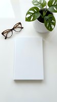 Minimalist workspace with blank notebook, tortoiseshell glasses and monstera plant on white desk. Clean office setup for productivity and creative work.